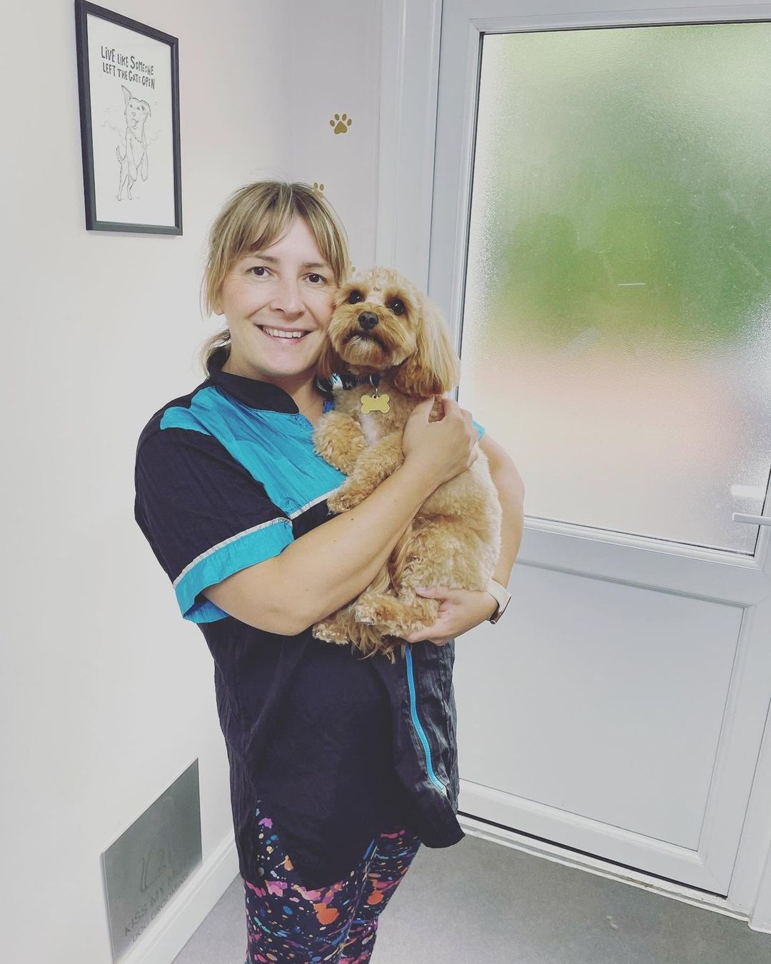Magda cuddling a cockapoo-type dog by the salon door at Kiss My Mutt, Stone