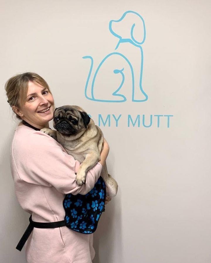 Magda holding a pug in front of the Kiss My Mutt wall logo at Kiss My Mutt, Stone